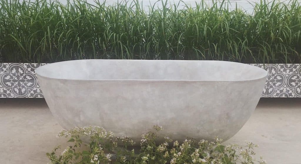 Bồn tắm đá nhân tạo solid surface màu xám nhạt với thiết kế mộc mạc, đặt trong không gian ngoài trời, kết hợp cây xanh và gạch hoa cổ điển, tạo nên không gian thư giãn gần gũi với thiên nhiên.
